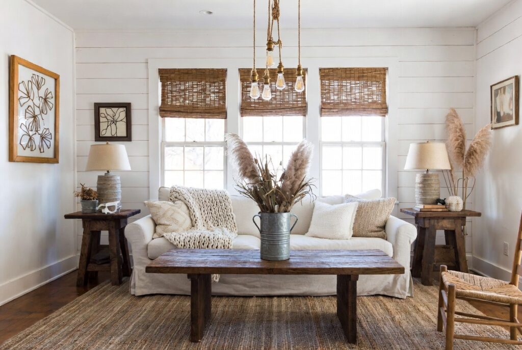 A cozy farmhouse living room featuring shiplap walls, a linen slipcovered sofa with chunky knit throw, reclaimed wood coffee table, large jute rug, galvanized metal vase with pampas grass, woven wood blinds, and layered warm Edison bulb lighting — illustrating farmhouse living room decor ideas for a warm and inviting homestead aesthetic.
